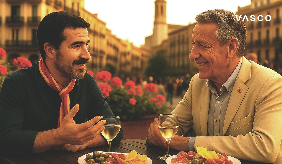 Dva muži si pochutnávají na drincích a tapas ve venkovní kavárně na Španělském náměstí a konverzují s květinami a architekturou v pozadí.