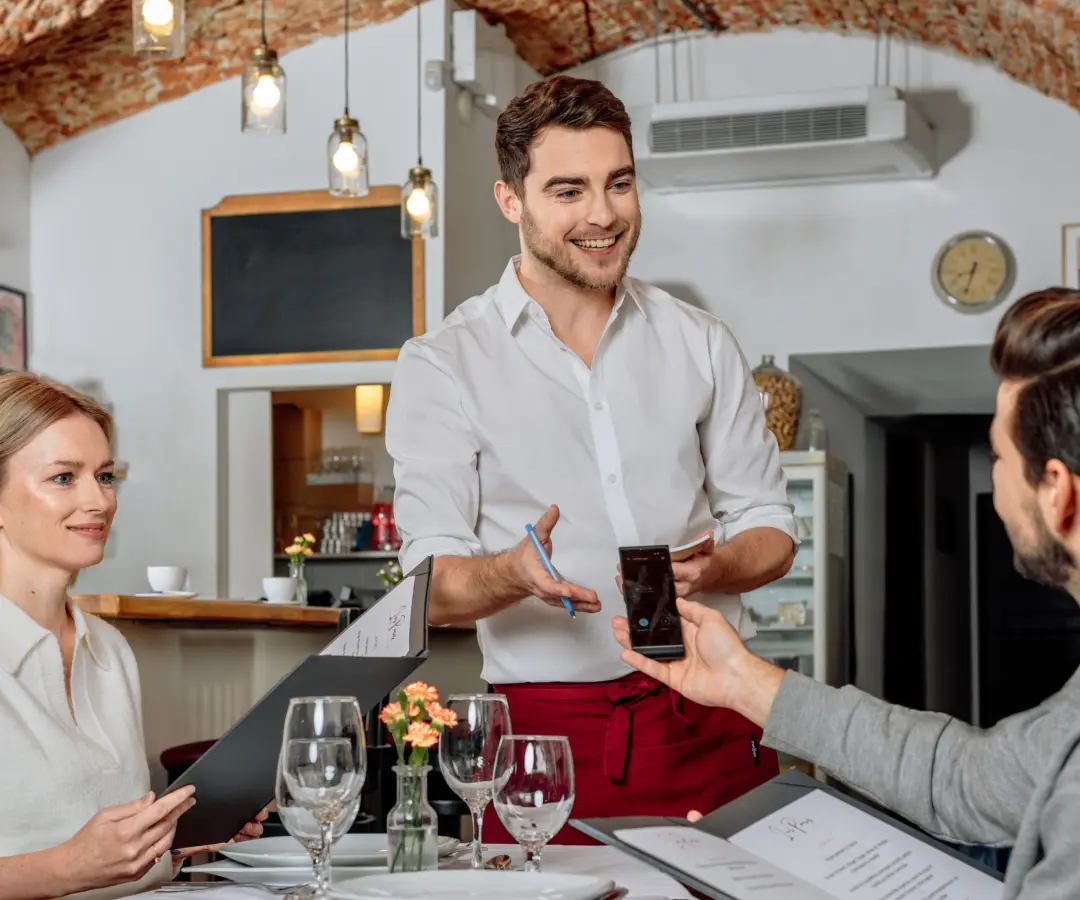 Usmívající se číšník stojí u stolu v restauraci, zatímco sedící mužský zákazník drží v ruce překladač. Vedle sedí zákaznice s jídelním lístkem. Stůl je prostřen sklenicemi, talíři a malou vázou s květinami.