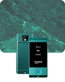 Prezentační fotografie produktu zachycující tyrkysové zařízení Vasco Translator ze dvou úhlů na pozadí tmavé texturované látky. Zařízení vlevo je zobrazeno zezadu, přičemž je zdůrazněn objektiv fotoaparátu s bleskem a reliéfní logo „VA“. Zařízení vpravo je zobrazeno zepředu s rozsvíceným displejem, na kterém se zobrazují pozdravy „Hello!“, „VA“, „¡Hola!“ a logo „VASCO Translator“. Kruhové tlačítko ve spodní části přední strany zařízení je orámováno modře svítícím světlem. Pozadí tvoří luxusní vzor zeleného mramoru se světlými žilkami.