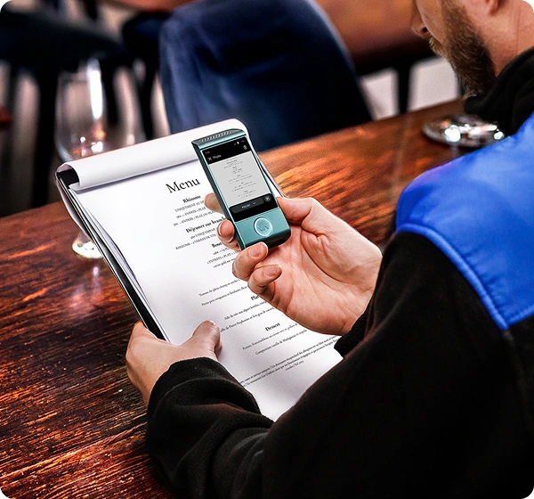 Záběr zblízka z nadhledu na osobu sedící u dřevěného stolu v restauraci, která si pomocí ručního elektronického překladatelského přístroje čte jídelní lístek. Osoba drží v levé ruce papírový jídelní lístek a pravou rukou nad ním drží malý překladatelský přístroj Vasco v barvě modrozelené. Na obrazovce zařízení se zobrazuje digitální verze jídelního lístku přeložená do polštiny. Osoba má na sobě černou bundu s jasně modrým panelem na rameni. Pozadí je jemně rozostřené a ukazuje útulný interiér restaurace. 