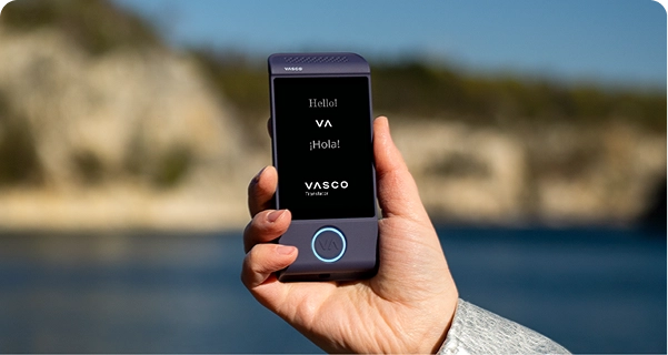 Detailní záběr ruky, která drží fialový elektronický překladatel Vasco, na pozadí jasného venkovního prostředí. Na displeji jsou zobrazeny pozdravy „Hello!“, „¡Hola!“ a logo „VASCO Translator“. Pozadí tvoří rozostřený pohled na slunnou pobřežní krajinu s modrou vodou a světlými skalnatými útesy pod jasnou oblohou. Osoba, která drží zařízení, má na sobě světle šedý nebo stříbrný texturovaný rukáv bundy. 