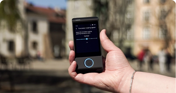 Detailní záběr na ruku osoby, která drží černý elektronický překladač značky Vasco, na pozadí rozmazaného venkovního záběru městského náměstí s historickými budovami. Na displeji zařízení je zobrazeno nastavení „Rychlost čtení překladu“ s posuvníkem nastaveným na „Normální“ a poznámkou: „Rychlost čtení překladu nastavíte posunutím posuvníku.“ Osoba držící zařízení má na ruce stříbrný náramek. Jasné denní světlo zvýrazňuje rozhraní obrazovky, díky čemuž jsou nastavení dobře viditelná. 
