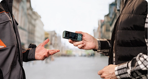 Muž v černé prošívané vestě přes kostkovanou košili drží elektronický překladač značky Vasco v barvě modrozelené vodorovně směrem k jiné osobě na městské ulici. Druhá osoba, oblečená v šedo-oranžové pracovní bundě, má nataženou ruku, jako by právě hovořila. Na displeji přístroje je vidět překladatelské rozhraní s textem. V pozadí je široký, rozmazaný pohled na ulici v evropském stylu s vysokými budovami pod jasnou, zataženou oblohou. 